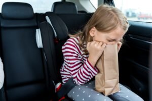 Little girl is driving in car. Kid is sick, feels bad and vomiting into paper bag. Traveling, riding on road in safe baby seats with child belts. Fun family trip, activity with parents